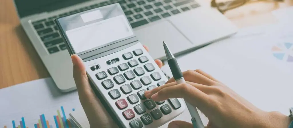 Hand bedient Taschenrechner, Unterlagen mit Diagrammen und Laptop auf Schreibtisch im Hintergrund