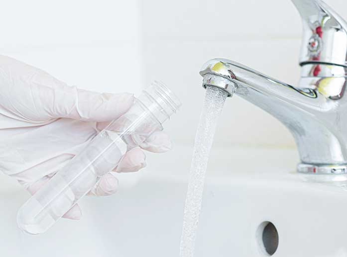 Hand in Plastikhandschuhen hält Probenbehälter unter fließenden Wasserhahn