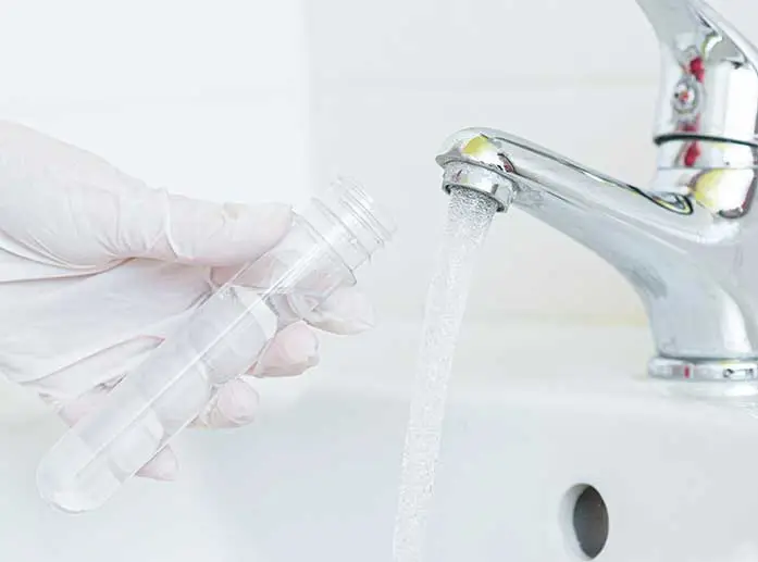 Hand in Plastikhandschuhen hält Probenbehälter unter fließenden Wasserhahn