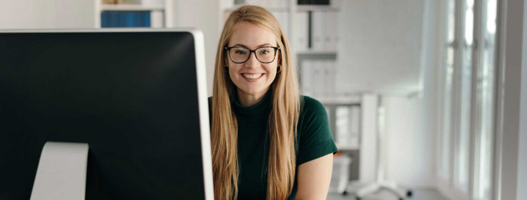 Frau lächelnd hinter Cumputerbildschirm am Schreibtisch