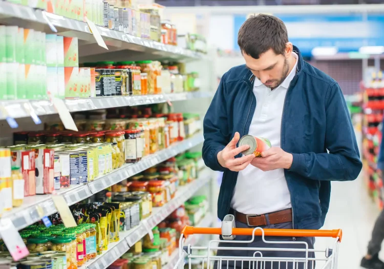 At the Supermarket: Handsome Man Uses Smartphone and Looks at Nutritional Value of the Canned Goods. He's Standing with Shopping Cart in Canned Goods Section.